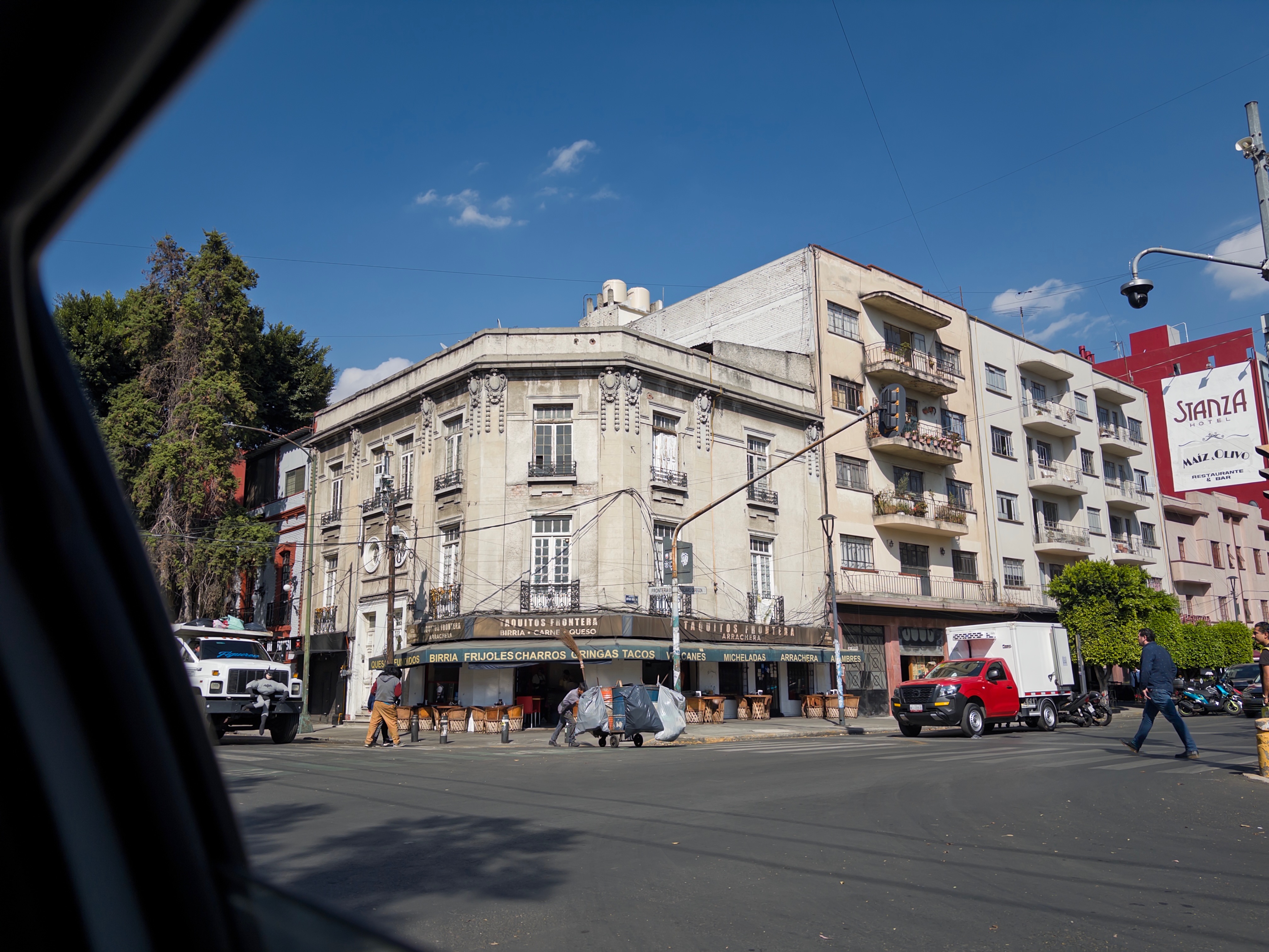A handsome early-20th-century corner building in Roma Norte, seen from across the street through the open window of a car on a clear morning. Stone detailing on the upper floors, ground-floor shops below, a Stanza hotel sign on the building to the right, pedestrians crossing the intersection.