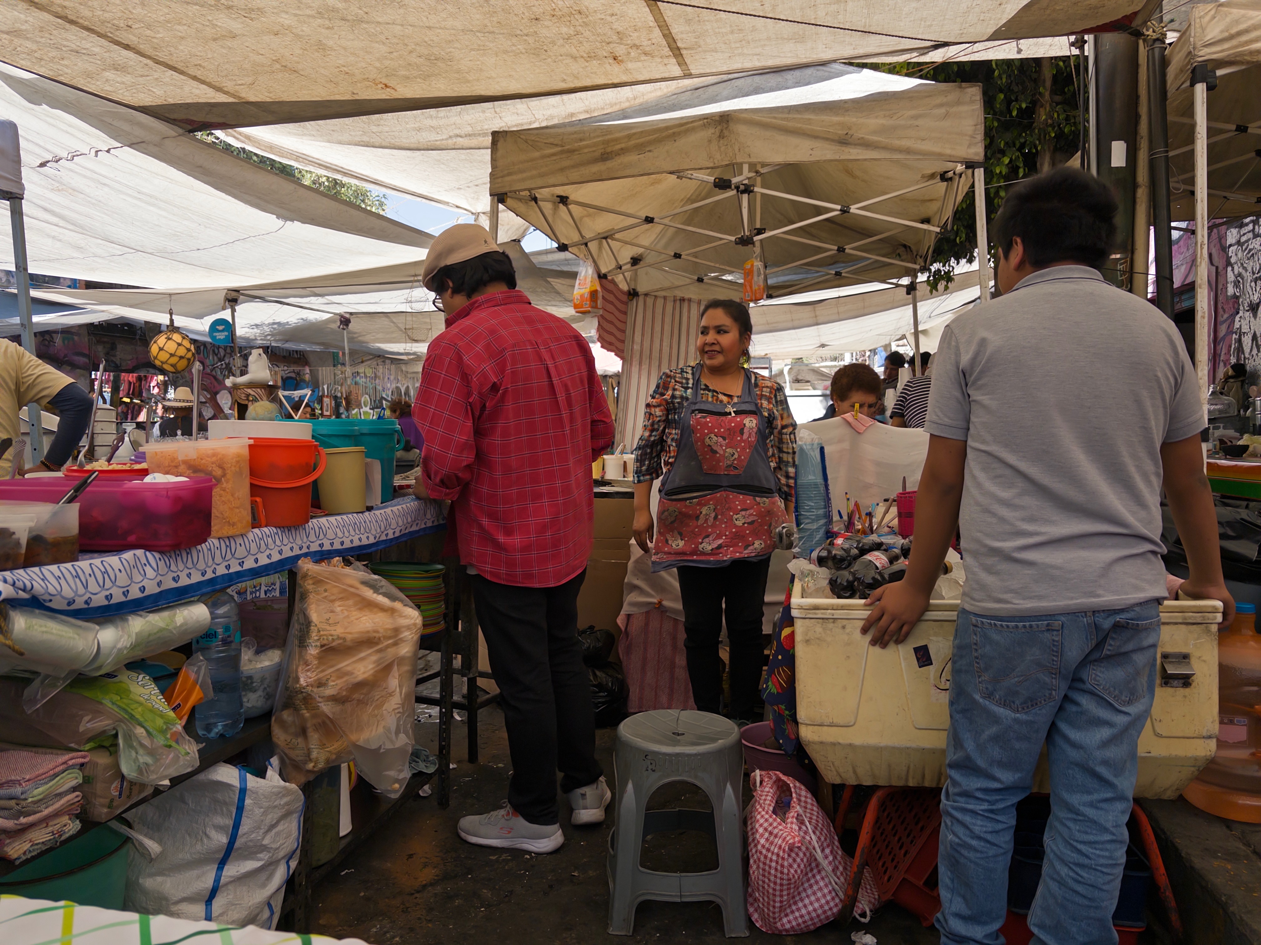 A woman in a red-heart apron stands behind her tianguis stall, greeting two shoppers across counters stacked with buckets, plastic bags, and household goods. White tarps stretch overhead.