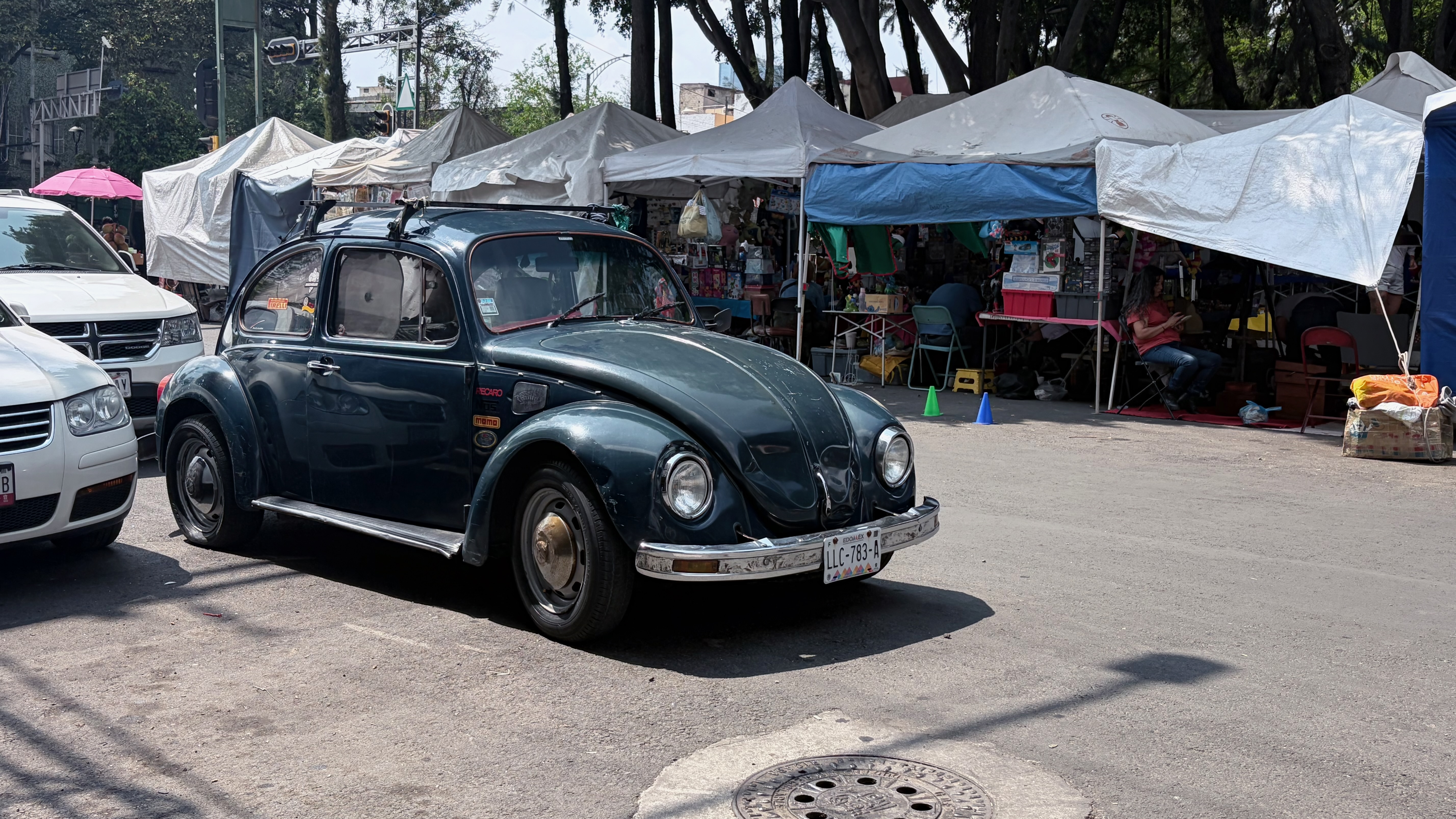 A black vintage Volkswagen Beetle parked at the curb beside a Sunday tianguis, with white and blue tarps stretched over the stalls behind it and trees lining the street.