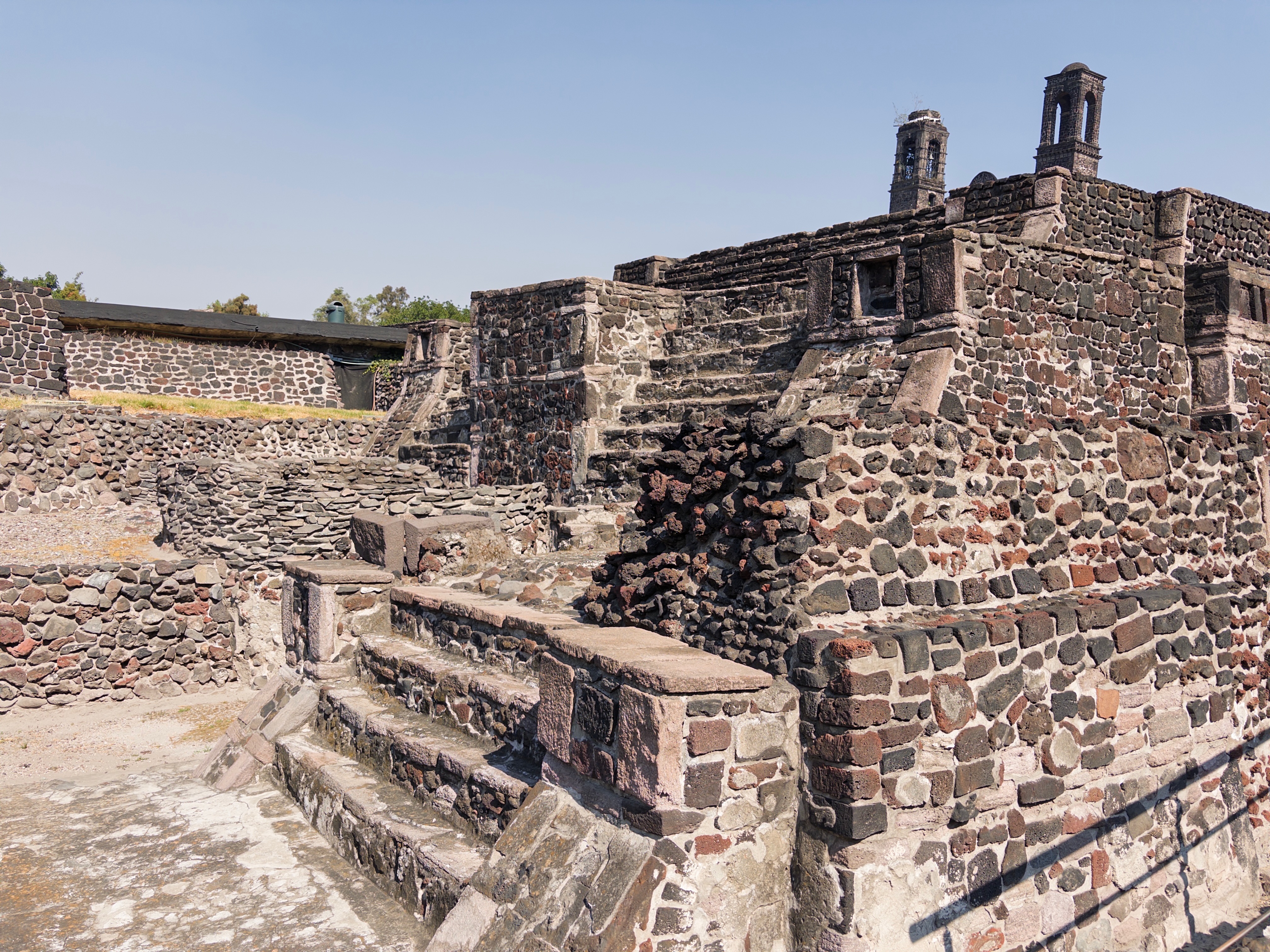 The pre-Hispanic stone ruins of Tlatelolco under a clear blue sky, weathered dark stone terraces and steps in the foreground, with the small twin bell towers of the colonial Iglesia de Santiago Tlatelolco rising behind the platform to the right.