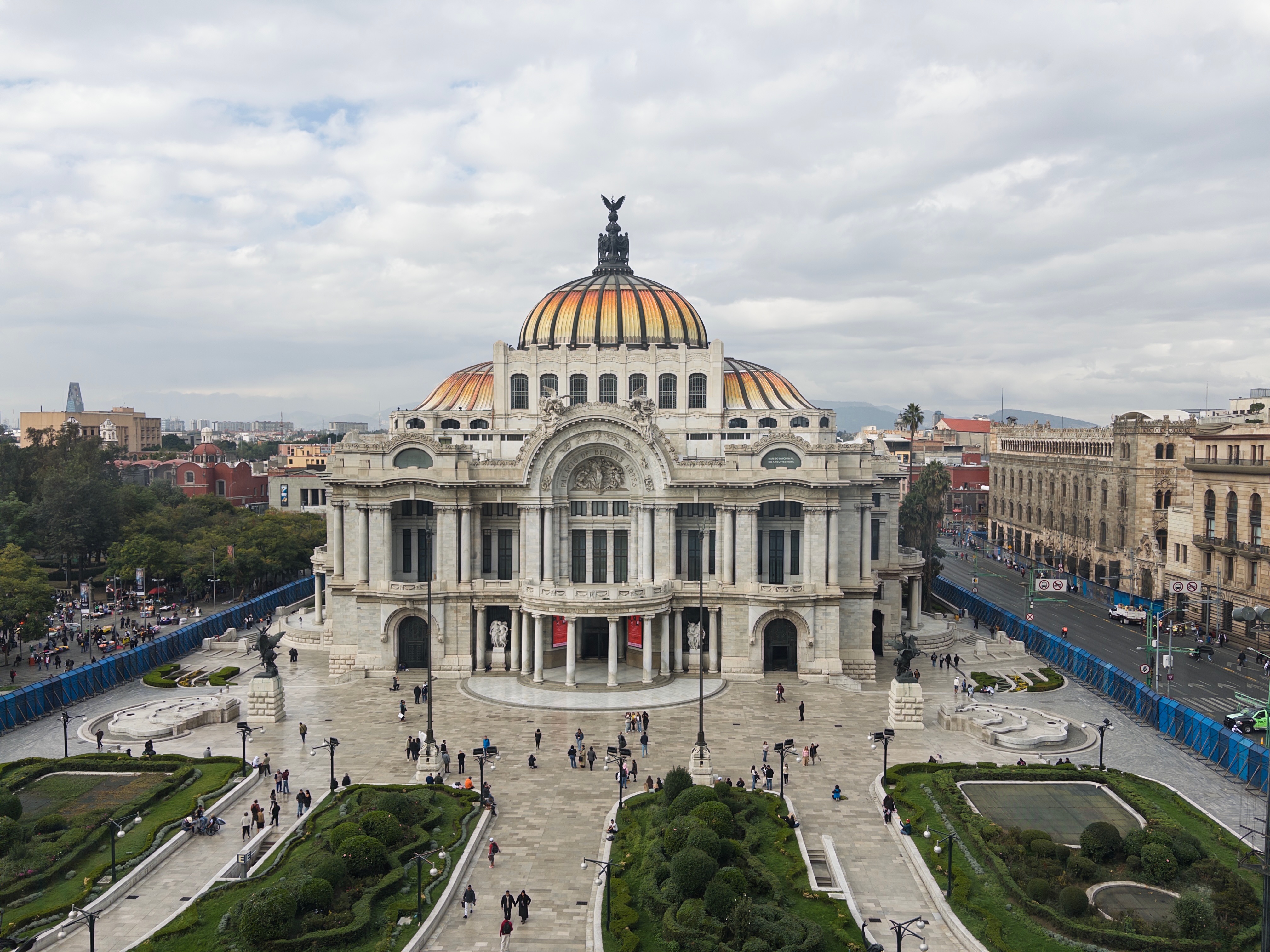 An elevated view of the Palacio de Bellas Artes in the Centro Histórico on an overcast day, its white marble neoclassical facade topped by a large Art Nouveau dome tiled in orange and gold with a bronze eagle at the peak. The surrounding plaza is filled with scattered pedestrians, trimmed hedges, and ornamental fountains, with blue construction barriers lining the adjacent avenues and the broader city skyline visible in the background.