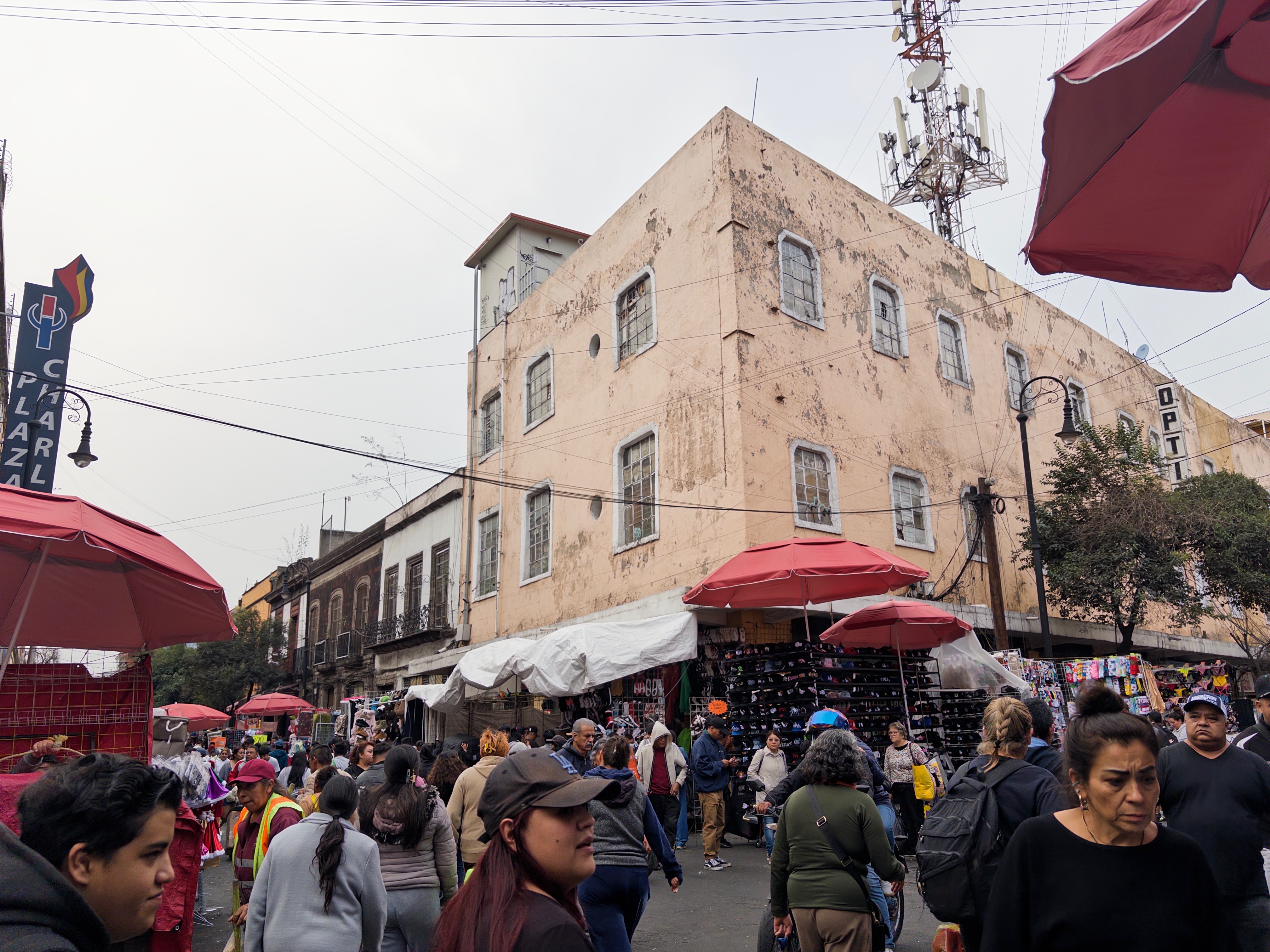 A busy outdoor street market in Centro Histórico, with crowds of shoppers moving past vendor stalls covered by red umbrellas and white tarps, shoe merchandise displayed on black grid racks, and a large weathered pink multi-story building behind them. A sign for Plaza Charl is visible on the left, a cell tower rises above the building, and the sky is overcast.
