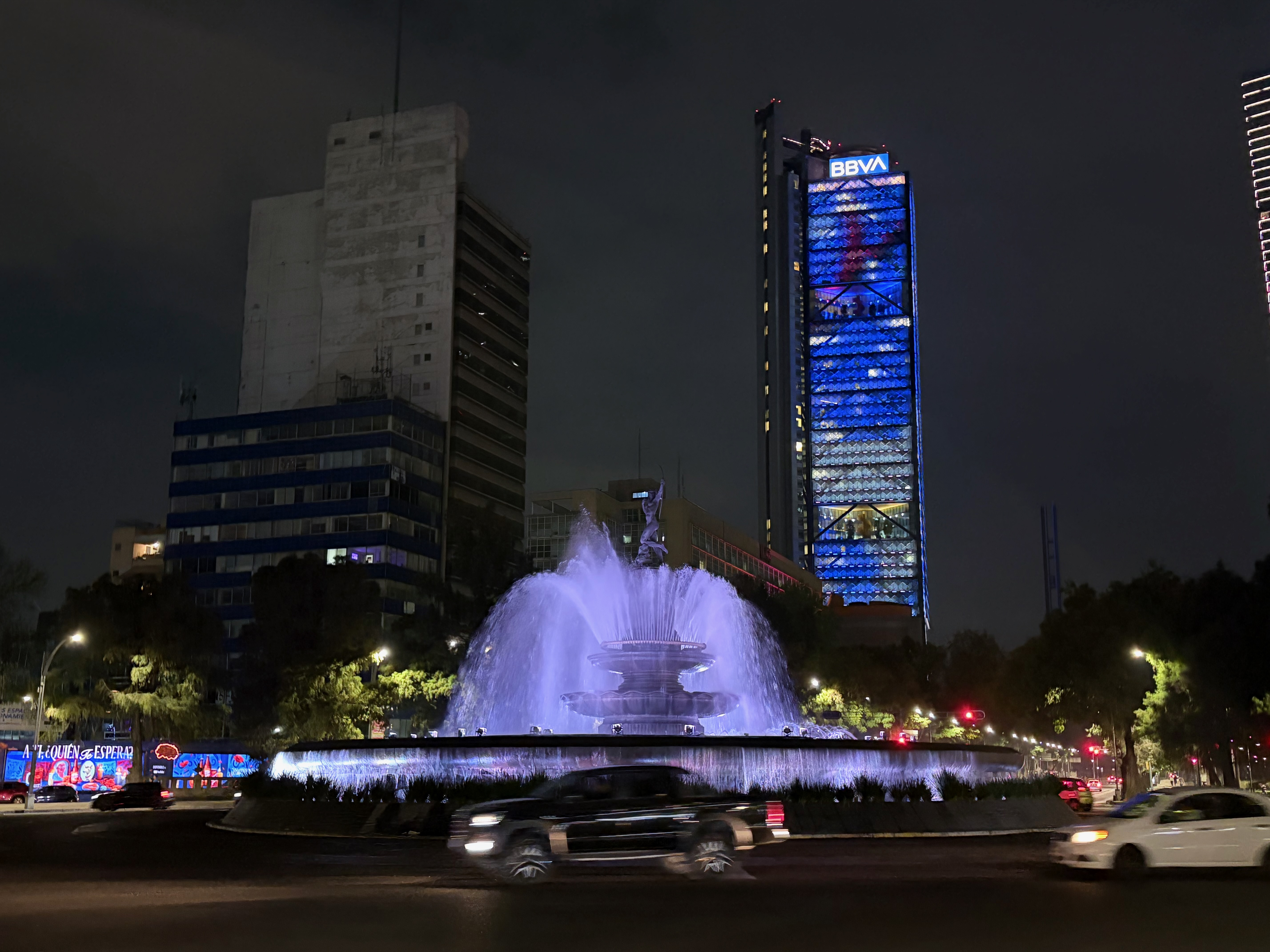 The Fuente de la Diana Cazadora roundabout fountain on Paseo de la Reforma, lit in blue-purple at night, with a statue of Diana at its center and water cascading from tiered basins. A dark SUV and a white sedan pass through the roundabout in the foreground, trees glow green under streetlights, and the BBVA tower—its facade illuminated in blue—rises in the background.