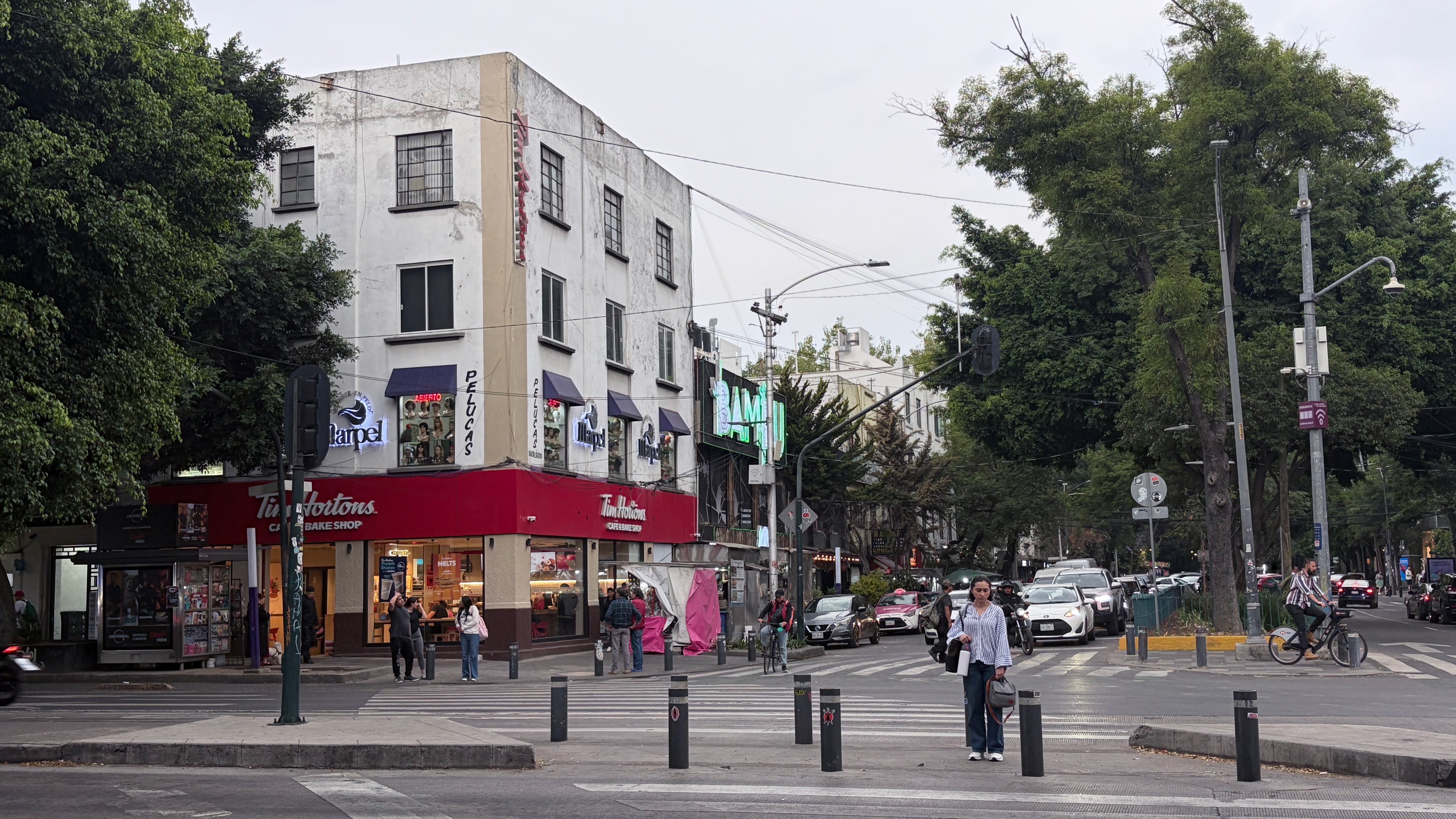 A busy pedestrian corner on Avenida Insurgentes in Condesa, with a red-awninged Tim Hortons café and bake shop occupying the ground floor of a weathered three-story white building. A wig shop called Marpel Pelucas sits above, small groups of people stand on the sidewalk, a woman in jeans and a striped blouse crosses the intersection in the foreground, cars queue along the boulevard under overcast midday skies, and large mature trees line both sides of the avenue.