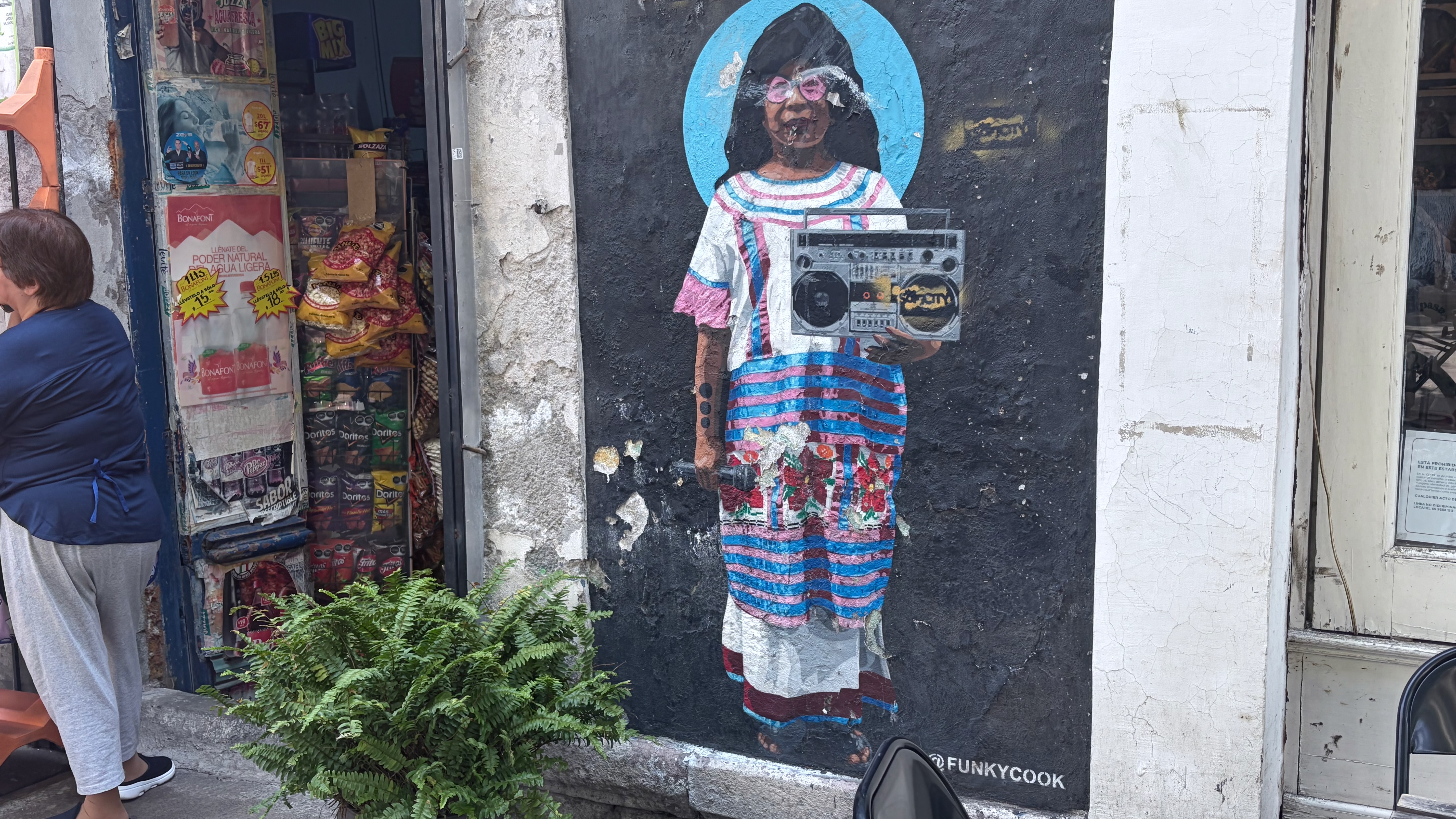 A street-art paste-up on a dark wall in Mexico City showing a young Indigenous woman in traditional striped and floral huipil dress, wearing pink heart-shaped sunglasses and holding a boombox, with a light-blue halo behind her head. The artwork is signed @FUNKYCOOK at the bottom right. To the left, a small convenience store displays chip bags, Bonafont water signage, and snack racks in its doorway; a fern grows from the sidewalk in front of the mural, and a pedestrian in a blue jacket and gray pants stands just outside the shop.