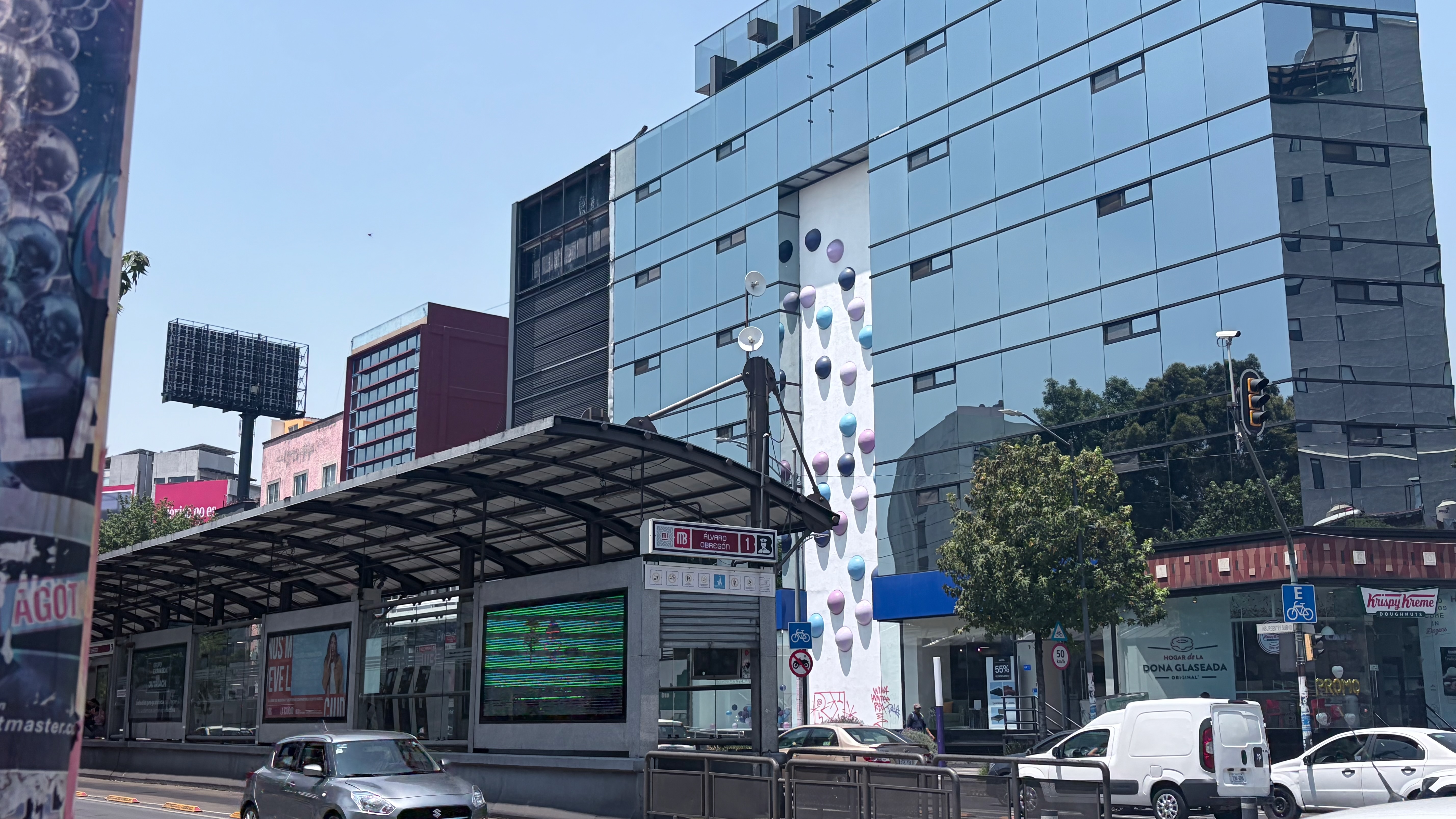 The Álvaro Obregón Metrobús station on a bright, clear afternoon along a busy boulevard in Roma Norte, with a covered platform, digital displays, and a station sign visible. Behind it rises a tall glass-curtain-wall office tower with a narrow white side wall decorated with large painted balloon shapes in purple, blue, and gray. A Krispy Kreme storefront and a Suzuki Swift in traffic appear in the foreground.