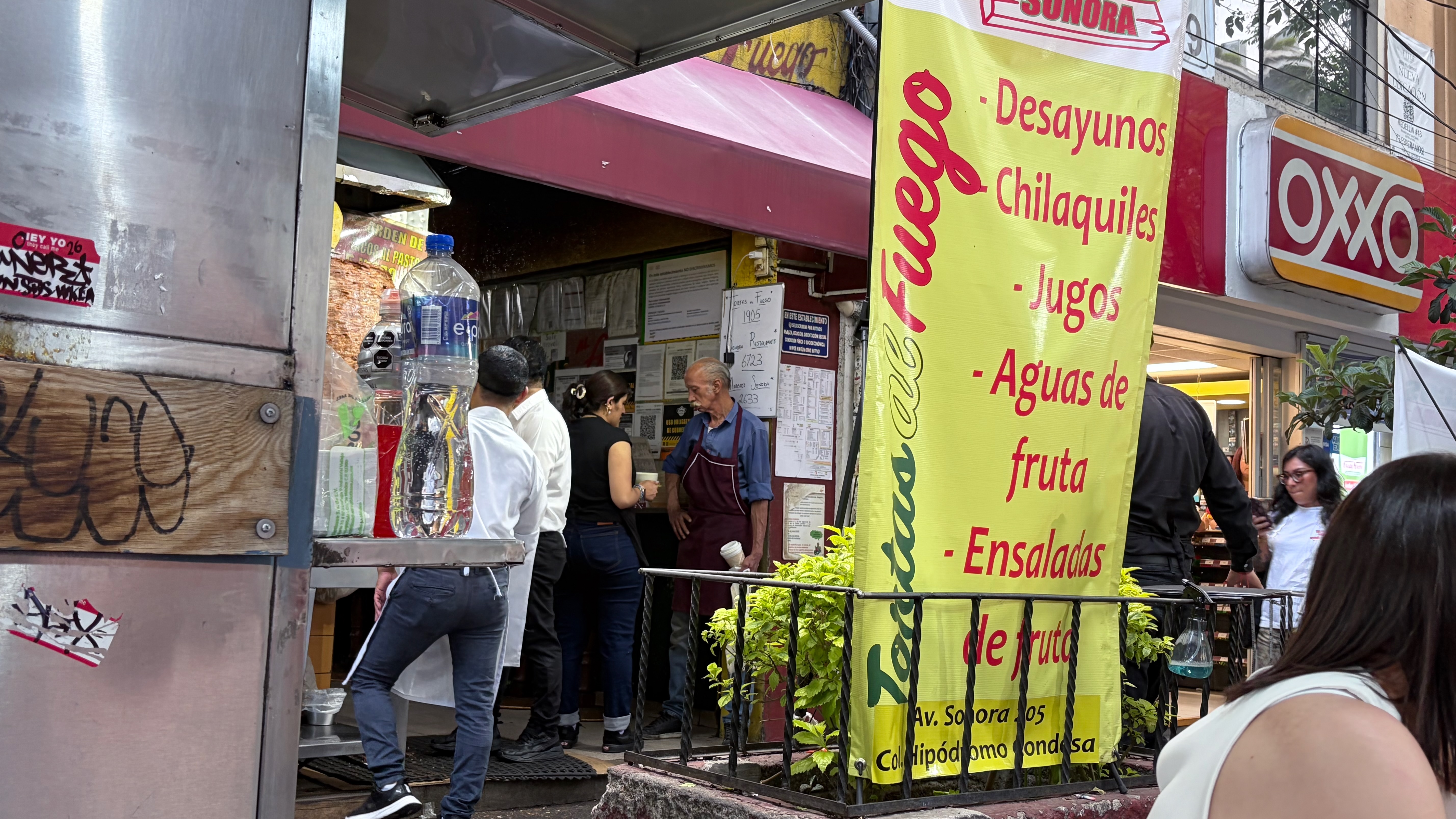 The entrance to Tacos al Fuego, a street-level restaurant at Av. Sonora 205 in Colonia Hipódromo Condesa, with a large yellow banner listing desayunos, chilaquiles, jugos, aguas de fruta, and ensaladas. Three people stand at the counter speaking with an older man in a burgundy apron, a large plastic water bottle sits on the counter to the left, and an OXXO convenience store is visible next door to the right.