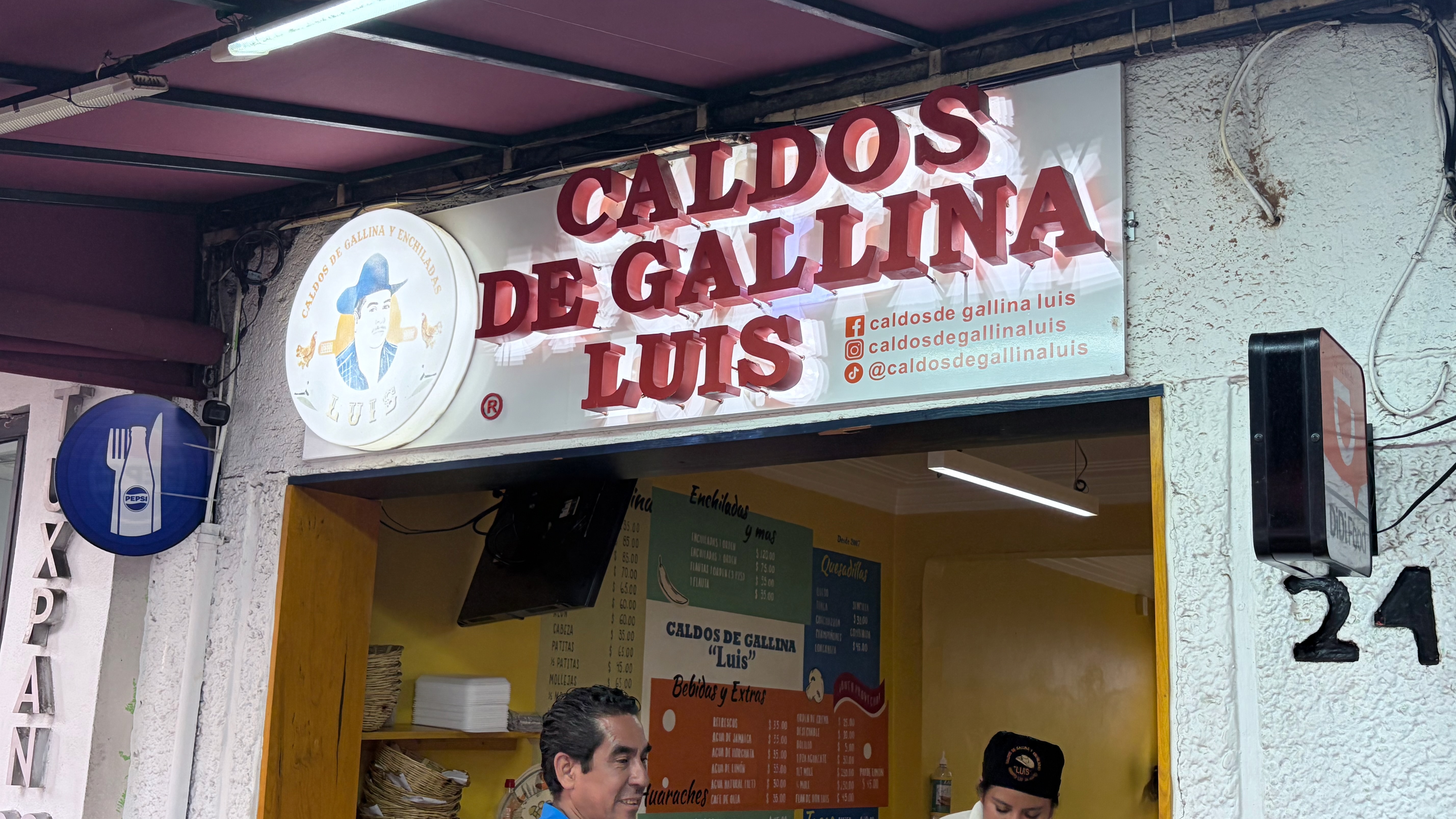 The storefront of Caldos de Gallina Luis, a small Mexican soup stall, with a large illuminated sign in red three-dimensional letters above the open counter. A logo medallion shows a mustachioed man in a blue hat. Two workers stand behind the counter, and menu boards listing caldos, enchiladas, quesadillas, and aguas are visible inside. A Pepsi sign hangs to the left.
