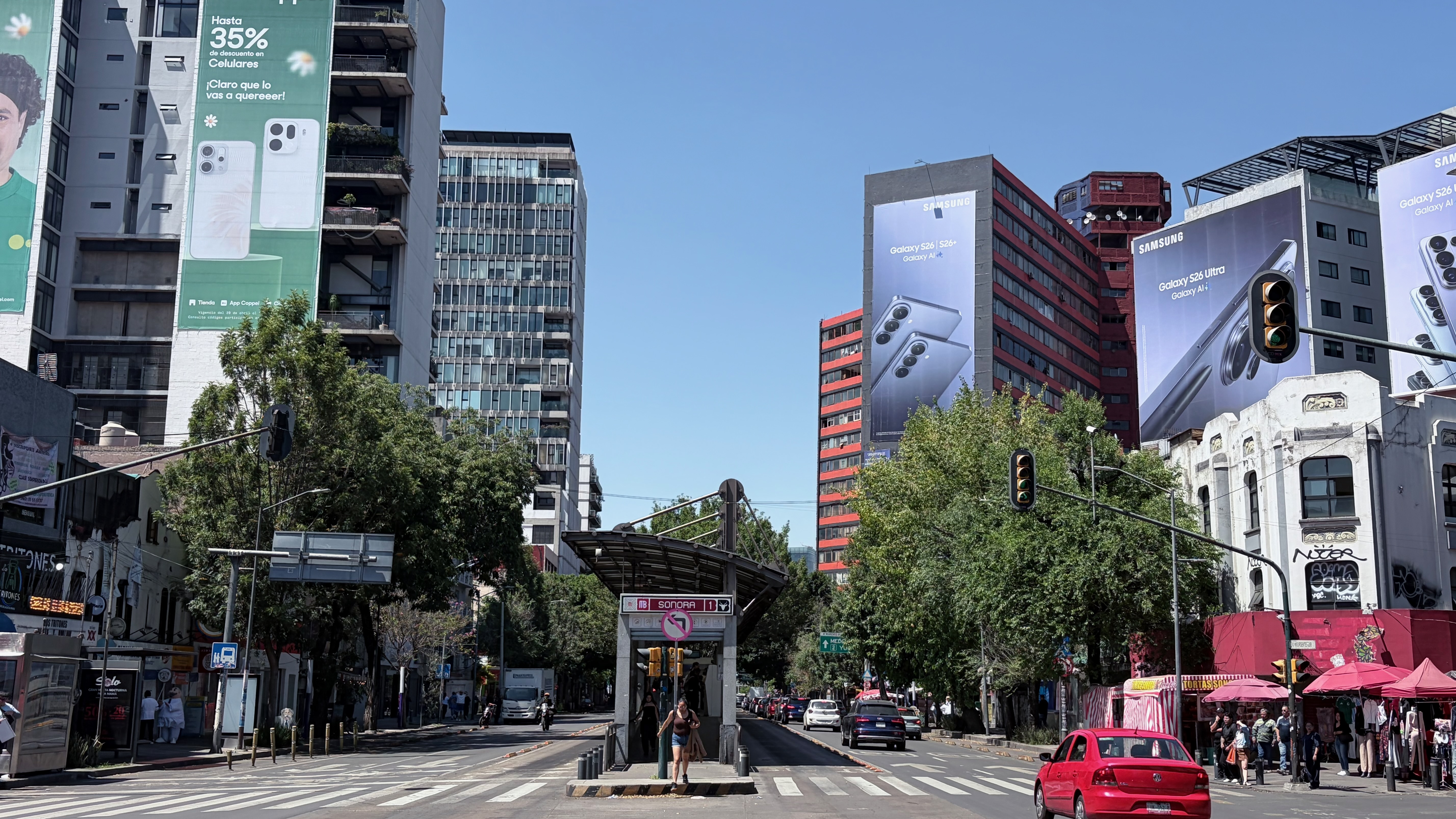 A busy urban intersection on a clear sunny day in Mexico City, centered on the entrance to Metro Sonora, a Line 8 station marked by a curved steel canopy and a red-and-white station sign. A woman in a summer dress crosses a zebra crossing in front of the entrance. Large Samsung Galaxy S26 billboard advertisements cover the faces of two buildings in the background, and a green Claro billboard advertising up to 35% off phones is mounted on a building to the left. A red sedan, a dark SUV, and several other cars occupy the street, with pedestrians and a pink-awninged market stall visible on the right sidewalk.