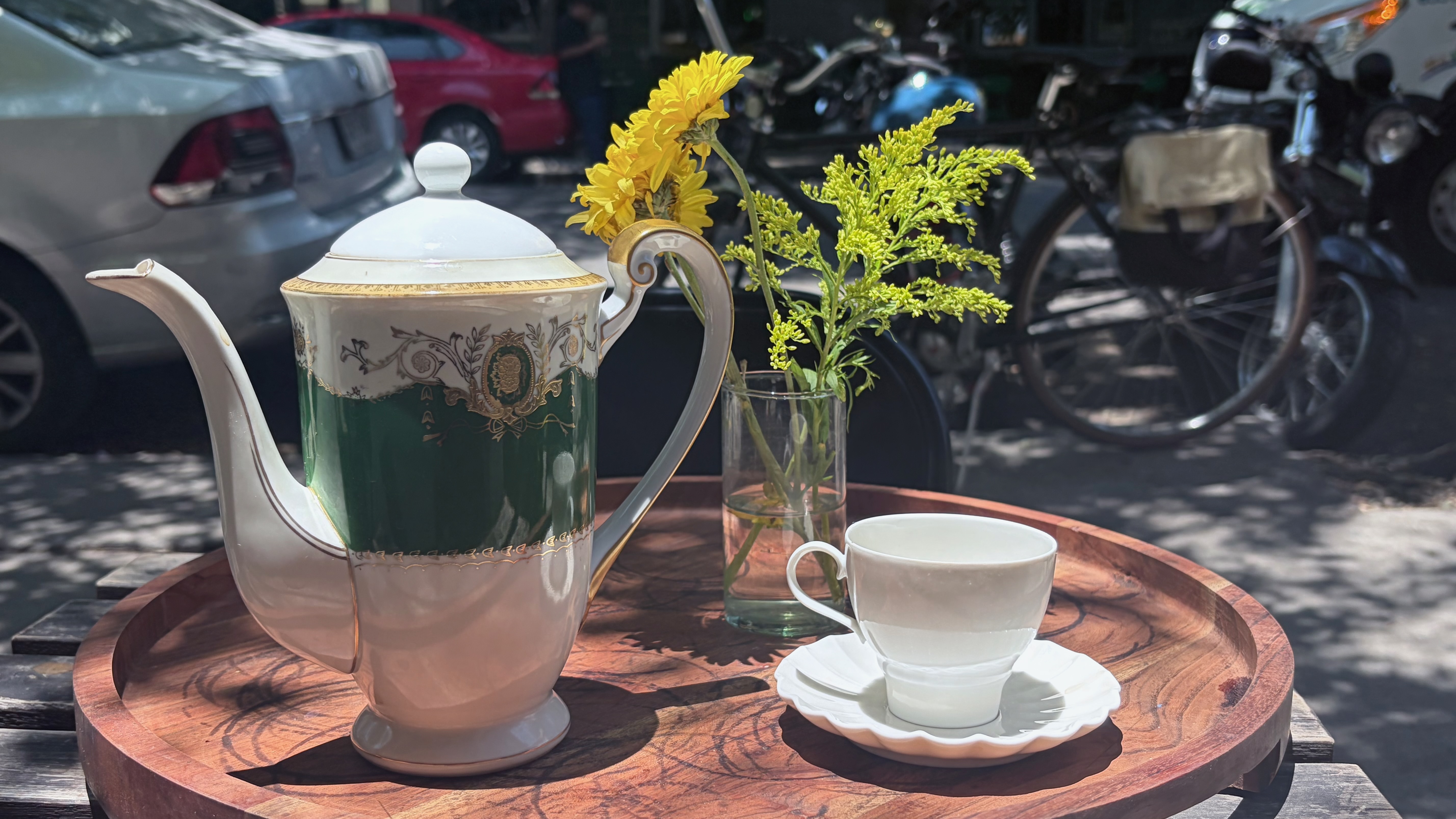 A white and green porcelain teapot with gold detailing and a matching white teacup on a saucer, arranged on a round wooden tray set on an outdoor table. A small glass vase holds yellow daisies and goldenrod. In the background, parked cars and motorcycles line a sunlit street.